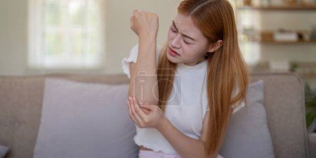 Young woman feeling elbow pain while sitting on sofa at home.