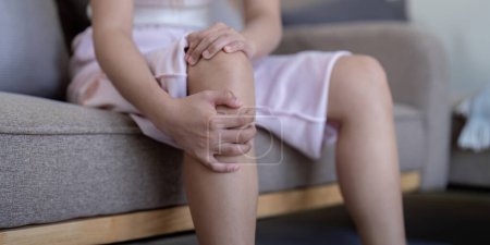 Foto de Young woman feeling knee pain while sitting on sofa at home. - Imagen libre de derechos