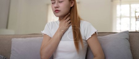 Young woman experiencing throat pain while sitting on a sofa at home.