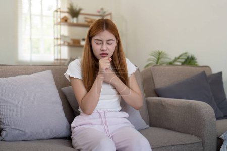 Young woman coughing while sitting on sofa at home.