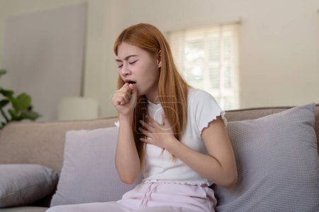 Young woman coughing while sitting on a sofa at home.