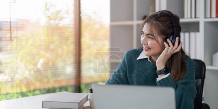 Photo for Young woman smiling while wearing headphones and using a laptop in a bright home office. - Royalty Free Image