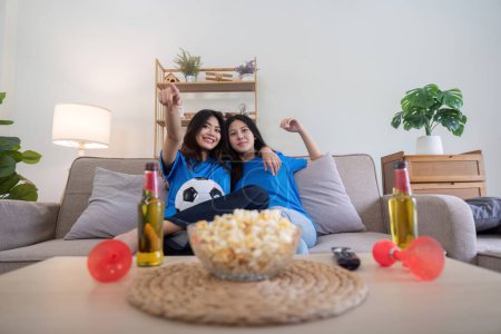 Foto de Pareja de lesbianas animando a Euro fútbol en casa con bebidas y palomitas de maíz. Concepto de orgullo LGBTQ, entusiasmo deportivo y celebración. - Imagen libre de derechos