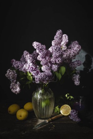 A bouquet of lilacs sits on a table with lemons.