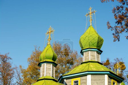 Foto de Histórica iglesia de madera de Juan el Teólogo en Pereiaslav, Ucrania. Cúpulas de cebolla verde con cruces doradas en la parte superior, colocadas contra un cielo azul y árboles de otoño - Imagen libre de derechos