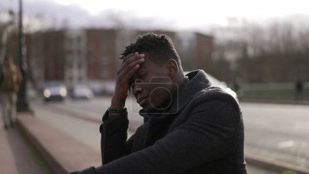 Foto de Anxious young black man suffering emotional pain sitting on sidewalk in street - Imagen libre de derechos