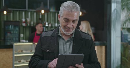 Téléchargez les photos : Portrait d'un entrepreneur mature tenant une tablette debout au café souriant à la caméra - en image libre de droit