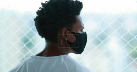 Téléchargez les photos : Black African man standing by window looking outdoors wearing epidemic mask prevention. person staying at home - en image libre de droit
