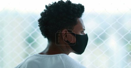 Téléchargez les photos : Black African man standing by window looking outdoors wearing epidemic mask prevention. person staying at home - en image libre de droit