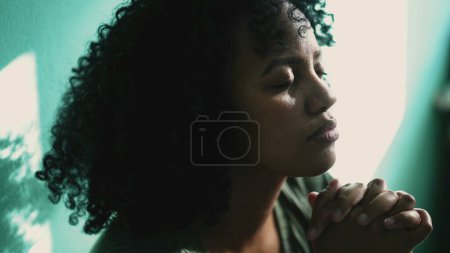Téléchargez les photos : Une jeune femme brésilienne pleine d'espoir priant à la maison. Une personne sud-américaine africaine ayant la foi 2 - en image libre de droit
