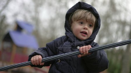 Téléchargez les photos : Toddler boy playing at playground gripping rope. Child wearing winter clothes plays outdoors holding cable - en image libre de droit