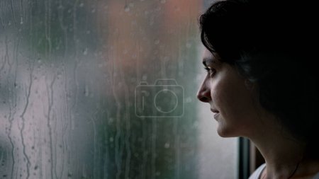 Mujer de pie junto a la ventana durante el día lluvioso mirando afuera viendo la lluvia