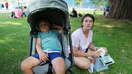 Foto de Child asleep inside stroller at park with mother sitting at grass. Family weekend lifestyle moment. Little boy napping - Imagen libre de derechos