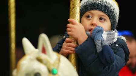 Téléchargez les photos : Un enfant heureux chevauchant carrousel à déplacer joyeux tour. Petit garçon s'amusant au parc d'attractions pendant la saison d'hiver. Fêtes de Noël - en image libre de droit