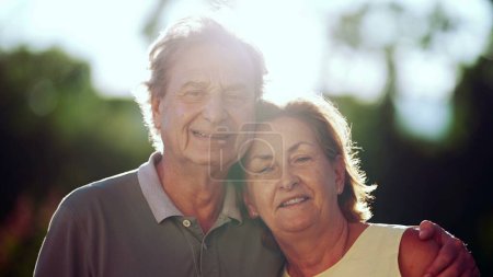 Seniorenpaar lächelt im Park in die Kamera. Porträtgesichter des Ehemanns mit Arm um die ältere Frau. Gegenlicht