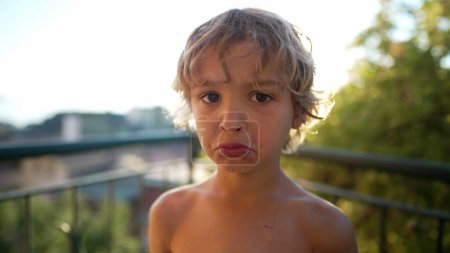 Téléchargez les photos : Portrait d'un jeune garçon avec une expression triste. Émotion sourde d'un petit garçon debout dehors regardant la caméra. Enfant malheureux - en image libre de droit