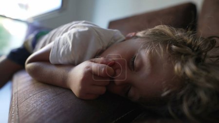 Téléchargez les photos : Petit garçon endormi piquant du nez alors qu'il était allongé sur le canapé. Enfant endormi sur le canapé mettant doigt à l'intérieur du nez - en image libre de droit
