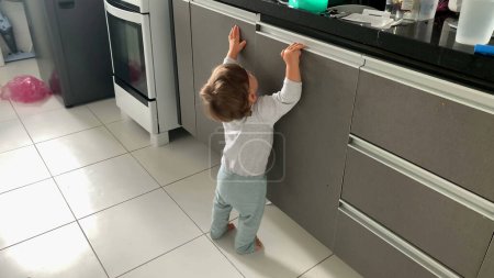 Foto de Bebé niño abriendo gabinetes de cocina. infante en dedos de los pies puntiagudos que buscan objetos - Imagen libre de derechos