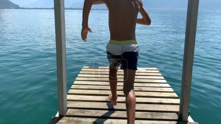 Téléchargez les photos : Jeune garçon courant et éclaboussant dans l'eau du lac. Le gamin court au quai en sautant et en plongeant dans l'eau - en image libre de droit