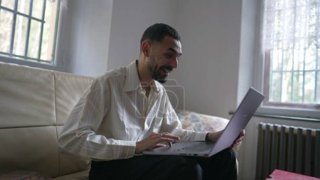 Foto de Un hombre emocionado feliz joven de Oriente Medio reaccionando positivamente al mensaje exitoso en línea mientras está sentado en el sofá delante de la computadora portátil - Imagen libre de derechos