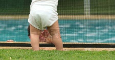 Foto de Cute baby standing by swimming pool fence security, infant looking at kids playing inside water - Imagen libre de derechos