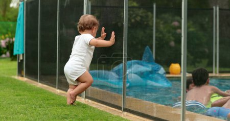 Foto de Bebé mirando a los niños jugar dentro del agua de la piscina durante el día sumemr. bebé apoyado en la valla de la piscina - Imagen libre de derechos