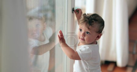 Téléchargez les photos : Baby standing by window wanting to leave. Cute infant toddler leaning on glass - en image libre de droit