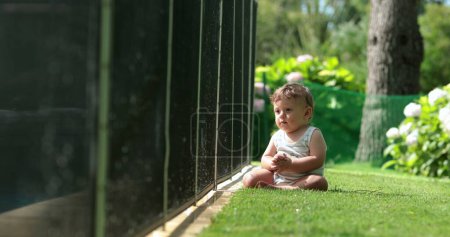 Foto de Contemplative cute baby seated outside on grass observing kids play at the pool - Imagen libre de derechos