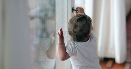 Téléchargez les photos : Baby toddler wanting to go outside holding window knob - en image libre de droit