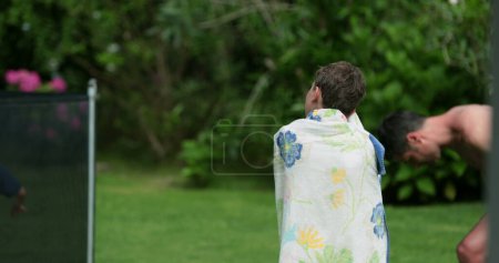 Téléchargez les photos : Enfant garçon enveloppé avec serviette après piscine marchant dans le jardin extérieur - en image libre de droit