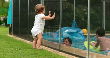 Téléchargez les photos : Mignon moment franc de bébé appuyé sur la clôture de la piscine voulant interagir avec les enfants à l'intérieur de la piscine - en image libre de droit
