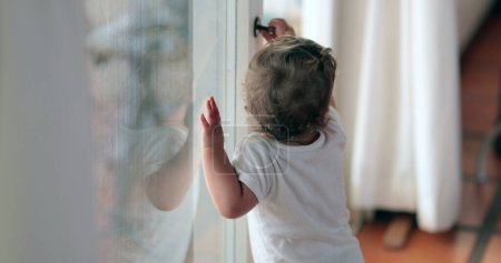 Téléchargez les photos : Baby toddler wanting to go outside holding window knob - en image libre de droit