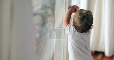 Téléchargez les photos : Bébé debout près de la fenêtre voulant partir. Bébé mignon tout-petit appuyé sur le verre - en image libre de droit