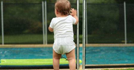 Photo for Baby leaning on swimming pool fence security. Infant in foreground while kids playing in background - Royalty Free Image