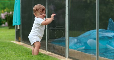 Foto de Bebé pequeño de pie junto a la valla de la piscina, prevención de ahogamiento, rejilla de protección - Imagen libre de derechos