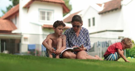Foto de Madre ayudando a su hijo con la tarea afuera. Mamá enseñando chico a leer en casa patio trasero - Imagen libre de derechos