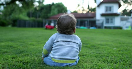 Téléchargez les photos : Bébé tout-petit suspendu à l'extérieur dans le jardin de jardin d'herbe de maison, maison résidentielle en arrière-plan - en image libre de droit