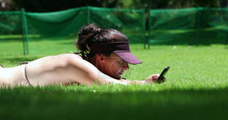 Téléchargez les photos : Femme franche utilisant un téléphone portable couché dehors sur l'herbe. Fille décontractée tapant sur smartphone en plein air - en image libre de droit