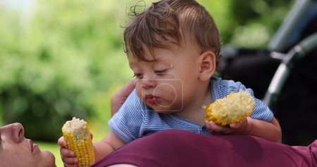 Foto de Lindo bebé comiendo mazorca de maíz afuera. Adorable bebé tomando un poco de bocadillo saludable al aire libre - Imagen libre de derechos