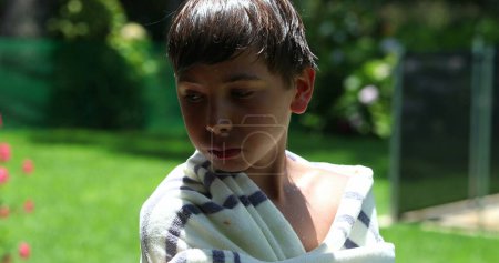 Téléchargez les photos : Enfant garçon mâchant de la nourriture enveloppée avec une serviette après la piscine pendant la journée d'été - en image libre de droit