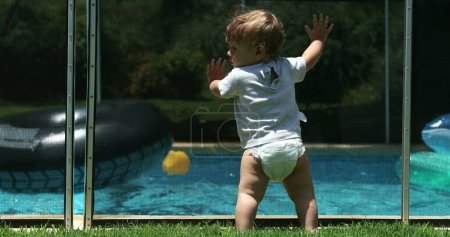 Photo for Cute infant baby leaning on swimming pool fence watching siblings play inside water - Royalty Free Image