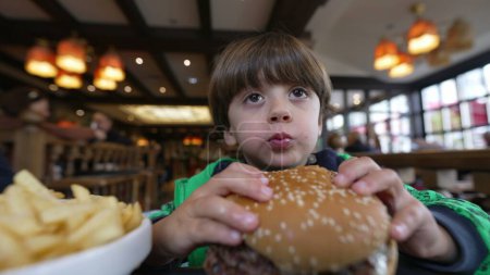 Téléchargez les photos : Un enfant attrape un hamburger et mange. Un petit garçon tenant un gros hamburger. Homme caucasien gosse prend une bouchée de délicieux aliments assis au restaurant à l'heure du déjeuner - en image libre de droit