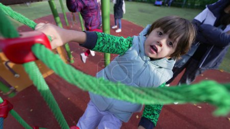 Téléchargez les photos : Corde d'escalade active pour enfants à l'extérieur de l'aire de jeux. Un garçon caucasien heureux faisant de l'exercice dehors grimpe. Développement de l'enfance style de vie - en image libre de droit