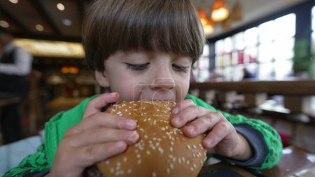 Photo for Young boy eating hamburger meal. Child taking a bite of burger food. Kid eats delicious lunch at restaurant diner - Royalty Free Image