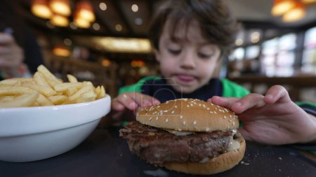Téléchargez les photos : Un petit garçon qui mange un hamburger assis au restaurant. Pensive enfant mange de la nourriture hamburger - en image libre de droit