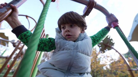 Téléchargez les photos : Enfant actif jouant à l'extérieur dans une aire de jeux. un mâle caucasien gosse exercice à l'extérieur holding sur métal bar - en image libre de droit
