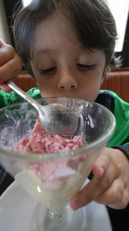 Téléchargez les photos : L'enfant mange une collation à la crème glacée avec une cuillère sur un bol en verre. Petit garçon assis au restaurant en train de manger un dessert en vidéo verticale - en image libre de droit
