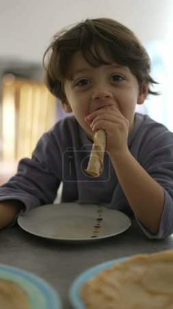 Foto de Un niño pequeño comiendo panqueques por la mañana usando pijamas. Alegre macho caucásico niño snacking desayuno comida en vertical vídeo - Imagen libre de derechos