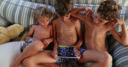 Téléchargez les photos : Enfants frères regarder du contenu en ligne sur tablette. Frères et sœurs ensemble regardant un film sur le canapé à la maison - en image libre de droit