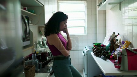Foto de Mujer joven introspectiva cándida de pie en la cocina del apartamento con expresión contemplativa. Persona pensativa pensando en la vida sola - Imagen libre de derechos